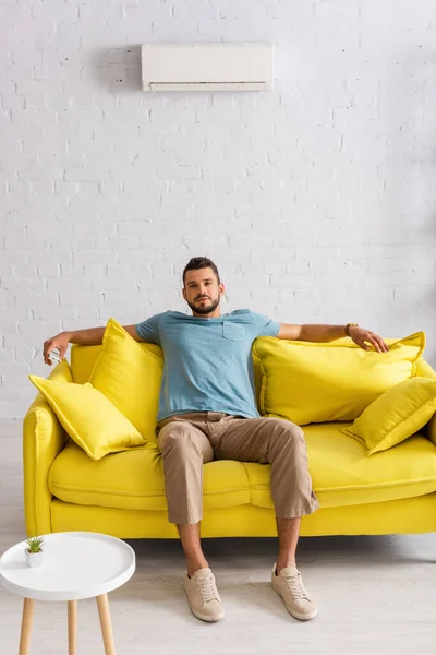 Selective focus of handsome man holding remote controller of air conditioner while looking at camera on sofa at home — Stock Photo