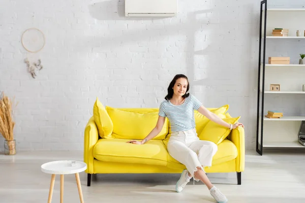 Mulher bonita olhando para a câmera enquanto sentado no sofá perto do controlador remoto de ar condicionado na mesa de café — Fotografia de Stock