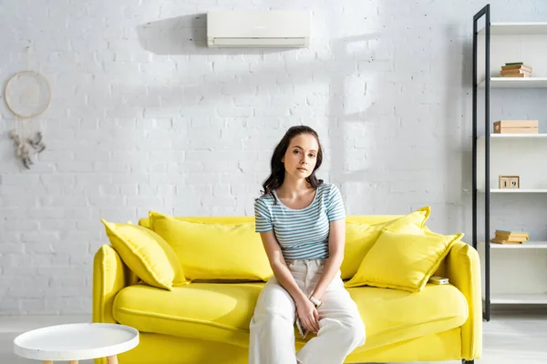 Attraktive Frau hält Fernbedienung der Klimaanlage, während sie auf der Couch sitzt — Stockfoto