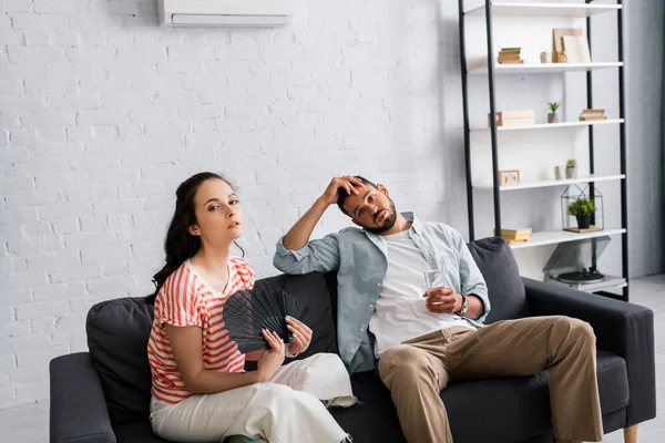 Pareja cansada con ventilador y vaso de agua mirando a la cámara mientras sufre de calor en casa - foto de stock