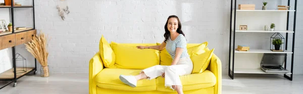 Imagen panorámica de la joven mujer sonriendo a la cámara mientras está sentada en el sofá amarillo en casa - foto de stock