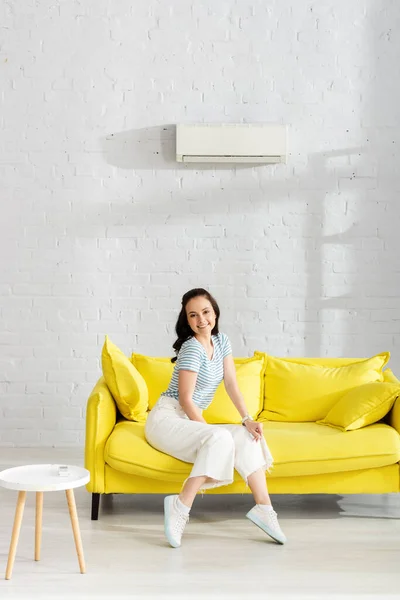 Menina atraente sorrindo para a câmera enquanto sentado no sofá perto do controlador remoto de ar condicionado na mesa de café em casa — Fotografia de Stock