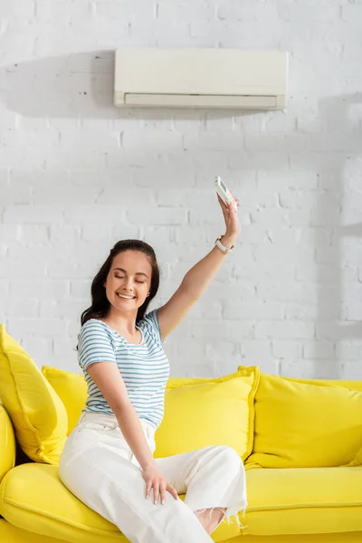 Sorrindo mulher segurando controlador remoto de ar condicionado no sofá na sala de estar — Fotografia de Stock