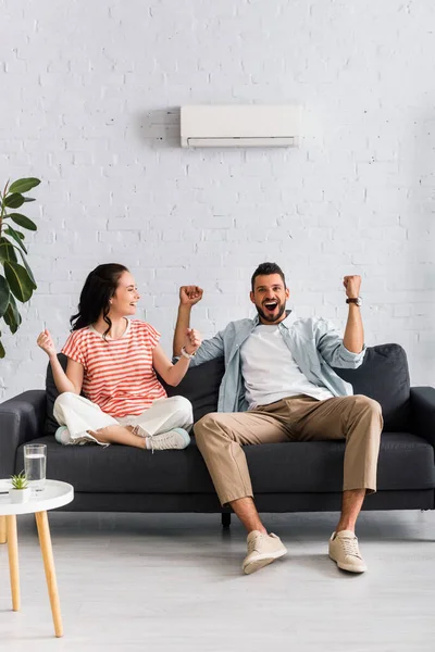 Coppia sorridente che mostra sì gesto mentre è seduto sotto il condizionatore d'aria sul divano — Foto stock