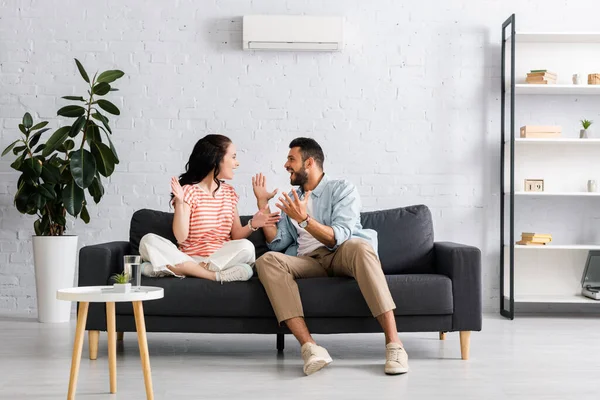 Positive couple smiling at each other while sitting on sofa under air conditioner at home — Stock Photo