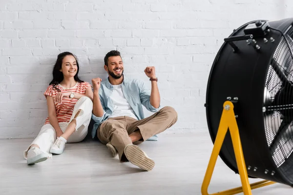 Selektiver Fokus eines positiven Paares, das auf dem Boden neben einem elektrischen Ventilator sitzt — Stockfoto