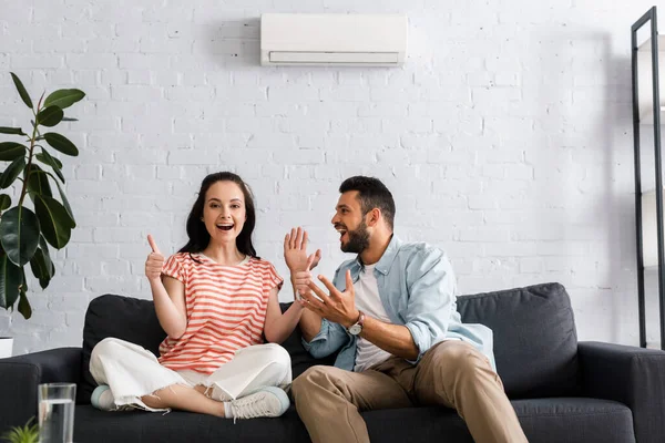 Selektiver Fokus einer lächelnden Frau, die im Wohnzimmer neben ihrem fröhlichen Freund Daumen nach oben zeigt — Stockfoto
