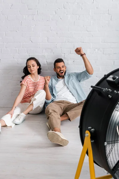 Selective focus of cheerful man showing yes gesture near girlfriend and electric fan on floor — Stock Photo