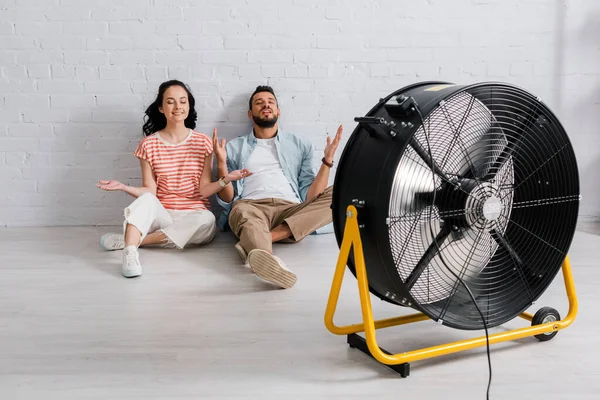 Счастливая пара сидит на полу рядом с электрическим вентилятором дома — стоковое фото