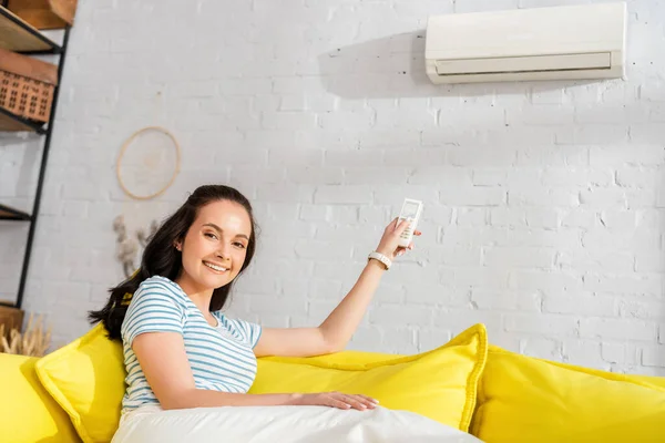 Mujer joven sonriendo a la cámara mientras cambia el aire acondicionado con mando a distancia en el sofá - foto de stock