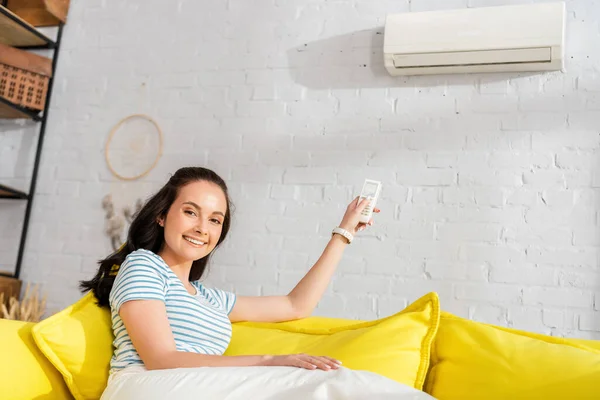 Jeune femme souriant à la caméra tout en commutant climatiseur avec télécommande sur canapé — Photo de stock