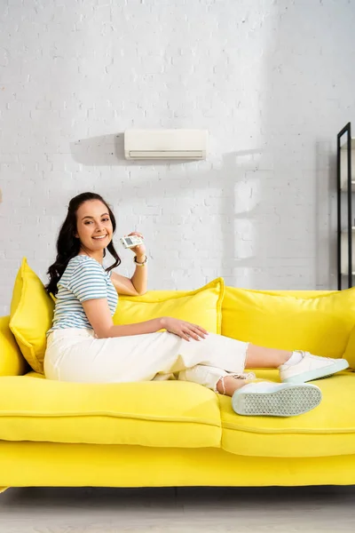 Bella donna sorridente alla macchina fotografica mentre tiene il telecomando del condizionatore d'aria in soggiorno — Foto stock