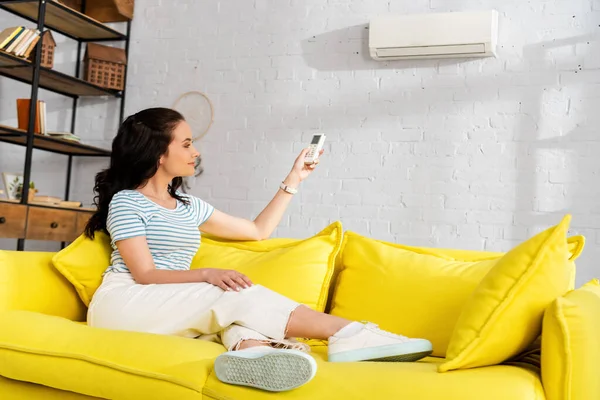 Schöne lächelnde Frau schaltet Klimaanlage, während sie zu Hause auf gelbem Sofa sitzt — Stockfoto