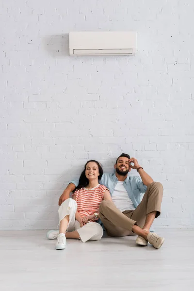 Casal jovem sorrindo para a câmera enquanto sentado sob ar condicionado no chão em casa — Fotografia de Stock