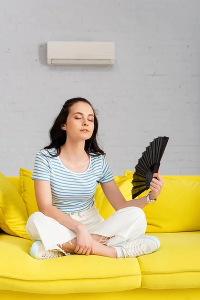 Jeune femme tenant ventilateur tout en souffrant de chaleur sur le canapé — Photo de stock