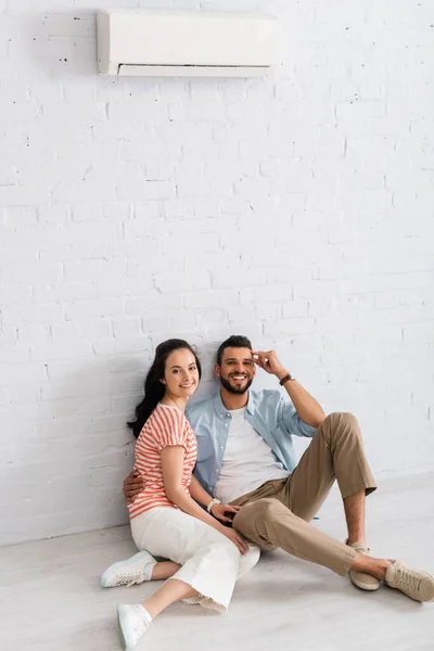 Barbudo hombre abrazando novia mientras está sentado en el suelo cerca de aire acondicionado en la pared en casa - foto de stock