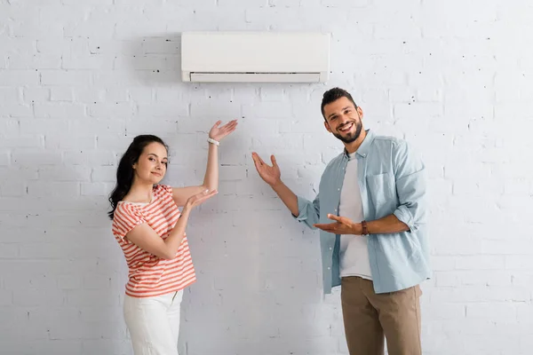 Couple souriant pointant des mains vers le climatiseur à la maison — Photo de stock