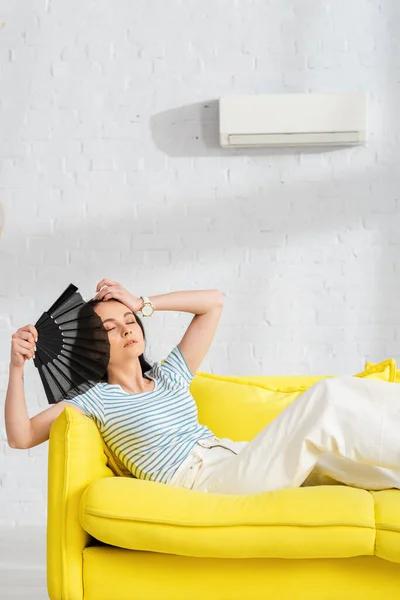 Mujer cansada sosteniendo ventilador mientras está sentado en el sofá con aire acondicionado en la pared en el fondo - foto de stock