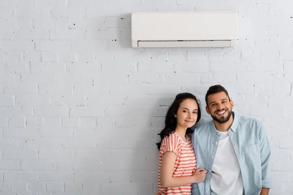 Casal jovem sorrindo para a câmera enquanto estava perto de ar condicionado na parede em casa — Fotografia de Stock