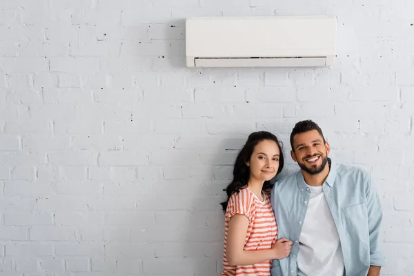 Pareja joven sonriendo a la cámara mientras está de pie cerca de aire acondicionado en la pared en casa - foto de stock