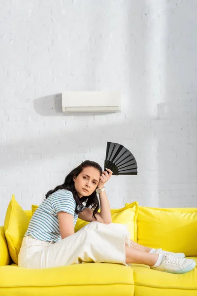 Seitenansicht einer traurigen Frau, die in die Kamera schaut, während sie einen Ventilator auf der heimischen Couch hält — Stockfoto