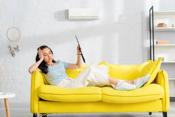 Selective focus of tired girl holding fan while suffering from heat on sofa in living room — Stock Photo