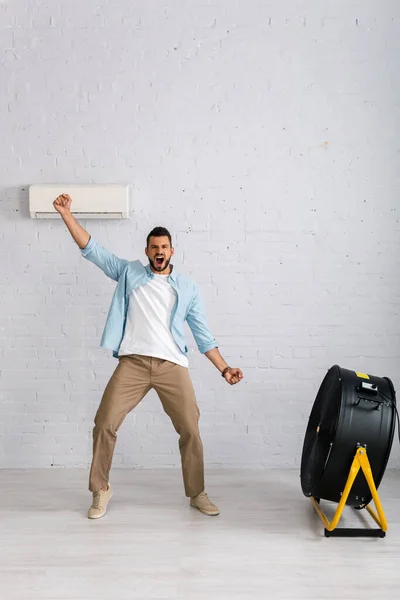 Hombre positivo mostrando sí gesto mientras está de pie cerca de ventilador eléctrico y aire acondicionado en casa - foto de stock