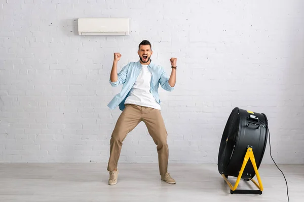 Homme positif montrant un geste oui tout en se tenant près du climatiseur et ventilateur électrique dans le salon — Photo de stock