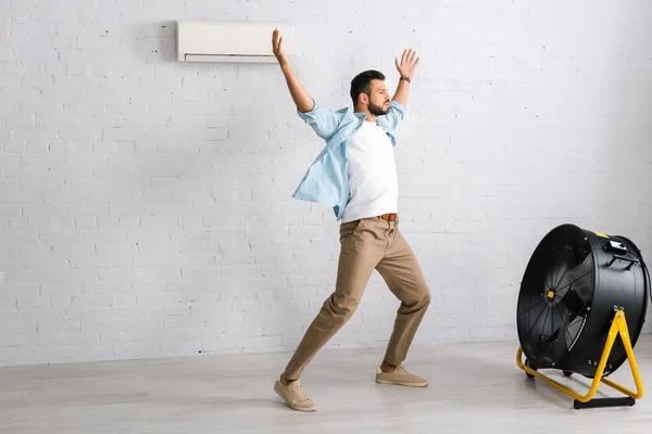 Hombre guapo de pie con las manos levantadas cerca de ventilador eléctrico y aire acondicionado en casa - foto de stock