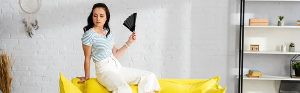 Panoramic crop of attractive girl holding fan on sofa in living room — Stock Photo