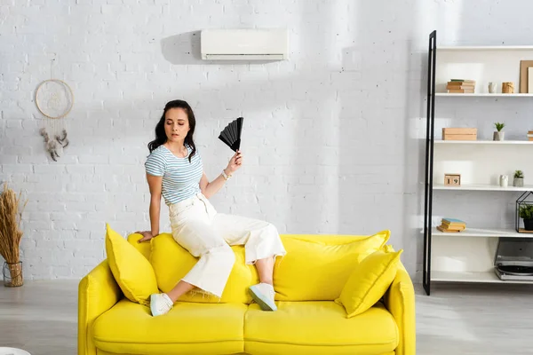 Schöne Frau hält Ventilator, während sie sich im Wohnzimmer heiß fühlt — Stockfoto