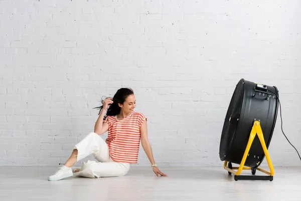 Attraktives lächelndes Mädchen, das auf dem Boden neben einem elektrischen Ventilator sitzt — Stockfoto