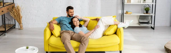 Recorte panorámico de mujer sonriente mirando a la cámara cerca de novio guapo en el sofá en casa - foto de stock