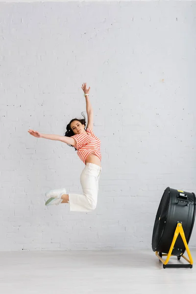 Junge Frau springt zu Hause mit erhobenen Händen neben Elektroventilator auf den Boden — Stockfoto