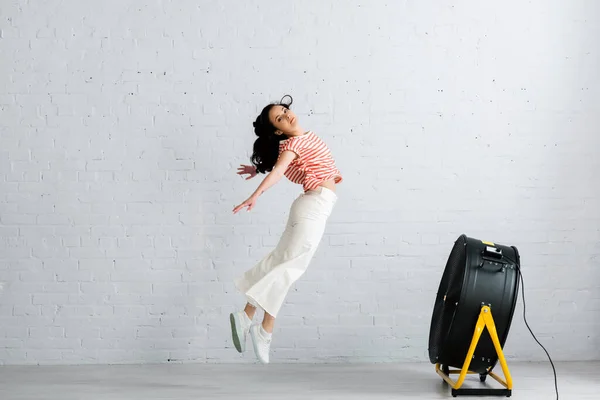 Вид сбоку привлекательной девушки, смотрящей на камеру во время прыжка рядом с электрическим вентилятором дома — стоковое фото