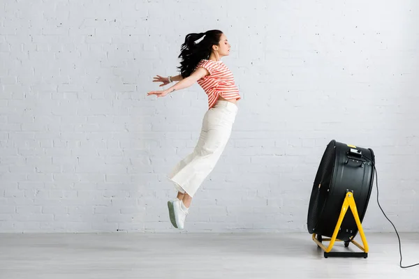 Vue latérale de la femme sautant près d'un ventilateur électrique à la maison — Photo de stock