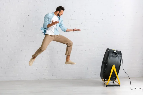 Schöner Mann springt zu Hause in der Nähe eines Elektroventilators auf den Boden — Stockfoto