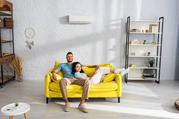 Beau homme souriant à la caméra près de belle petite amie couchée sur le canapé dans le salon — Photo de stock