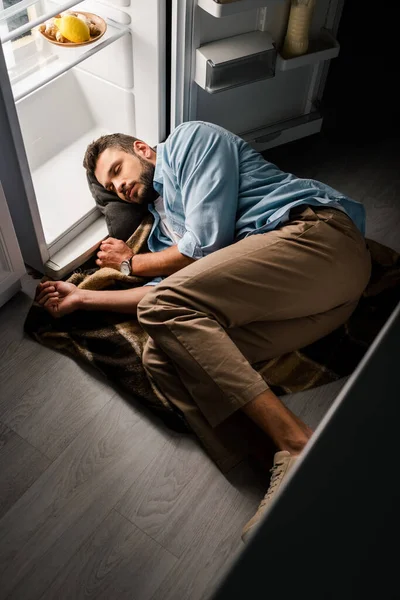 Вид под высоким углом на человека, спящего возле открытого холодильника на полу ночью — стоковое фото