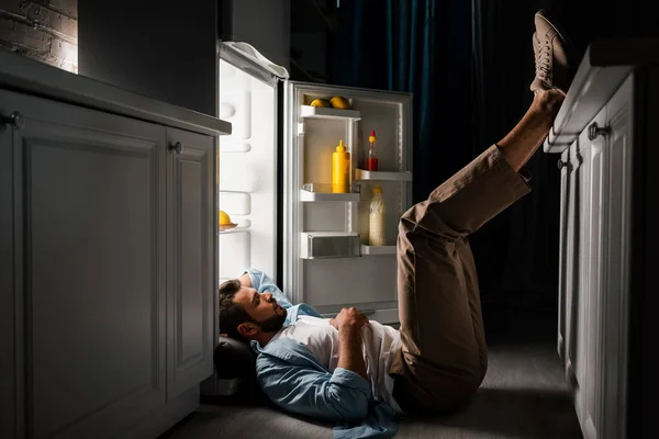 Vista laterale o uomo che dorme al piano vicino al frigorifero aperto di notte — Foto stock