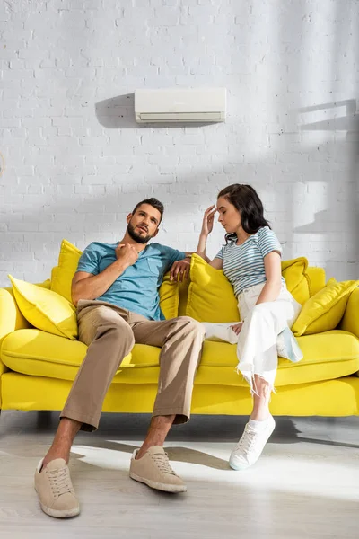 Pareja joven que sufre de calor con aire acondicionado en la pared en el fondo en la sala de estar - foto de stock