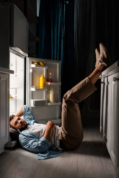 Vue latérale de l'homme regardant la caméra tout en étant couché sur le sol près du réfrigérateur ouvert la nuit — Photo de stock