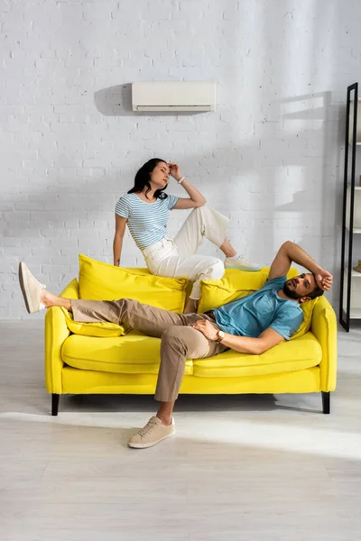 Young couple feeling hot while sitting on sofa with air conditioner on wall at background in living room — Stock Photo