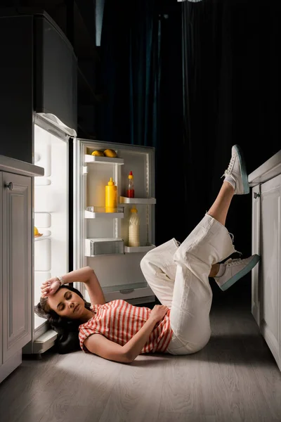 Bella donna sdraiata al piano vicino al frigorifero aperto in cucina di notte — Foto stock