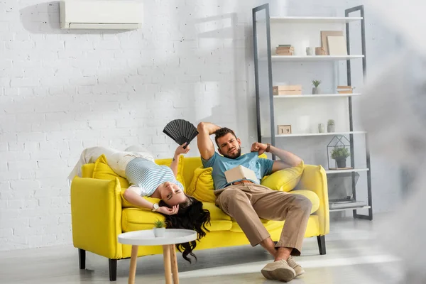 Selektiver Fokus eines erschöpften Paares mit Ventilator und Buch, das sich im Wohnzimmer heiß fühlt — Stockfoto