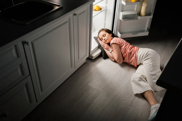 Высокоугольный вид привлекательной девушки, спящей на подушке возле открытого холодильника на кухне ночью — стоковое фото