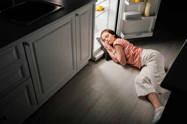 Vista de ángulo alto de chica atractiva durmiendo en la almohada cerca de nevera abierta en la cocina por la noche - foto de stock