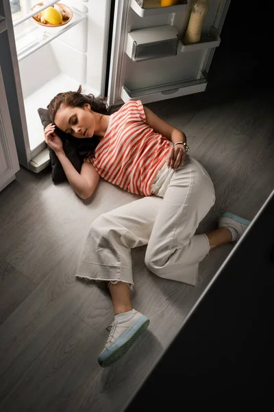 Vista de ángulo alto de la mujer joven durmiendo mientras está acostada cerca de refrigerador abierto en el suelo por la noche - foto de stock