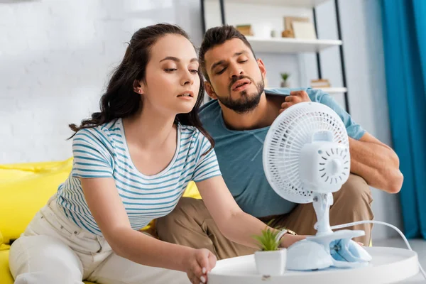 Selective focus of young couple sitting near electric fan on coffee table while suffering from heat — Stock Photo