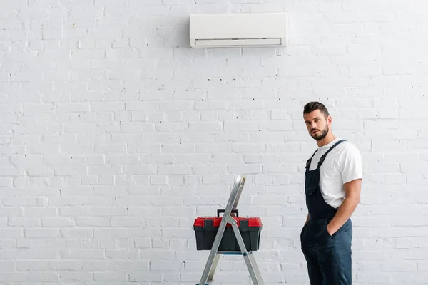 Schöner Arbeiter mit Händen in Hosentaschen und Blick auf die Kamera in der Nähe des Werkzeugkastens auf Leiter und Klimaanlage — Stockfoto